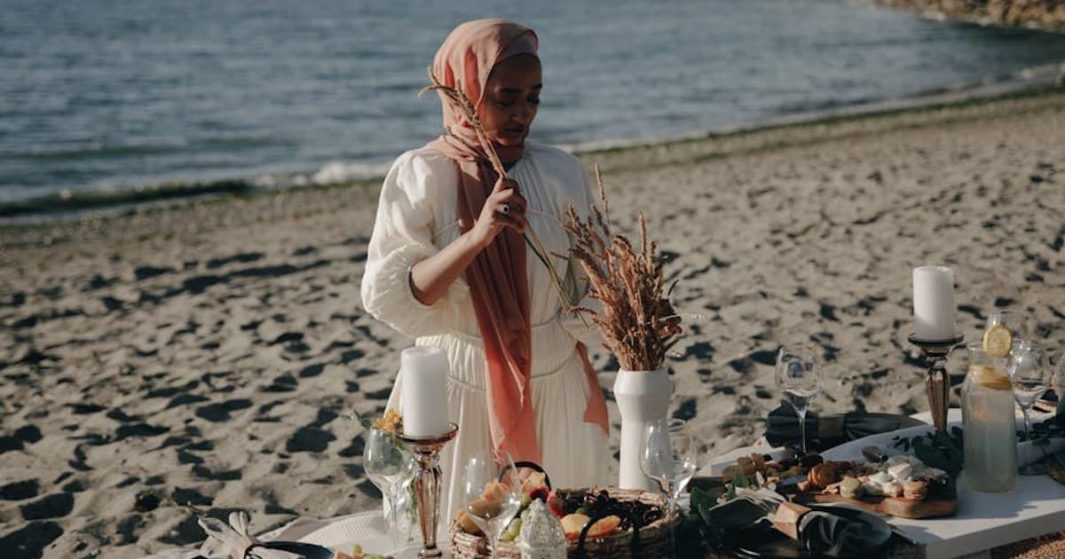 A woman in hijab arranging a dinner table by the beach on a sunny day.