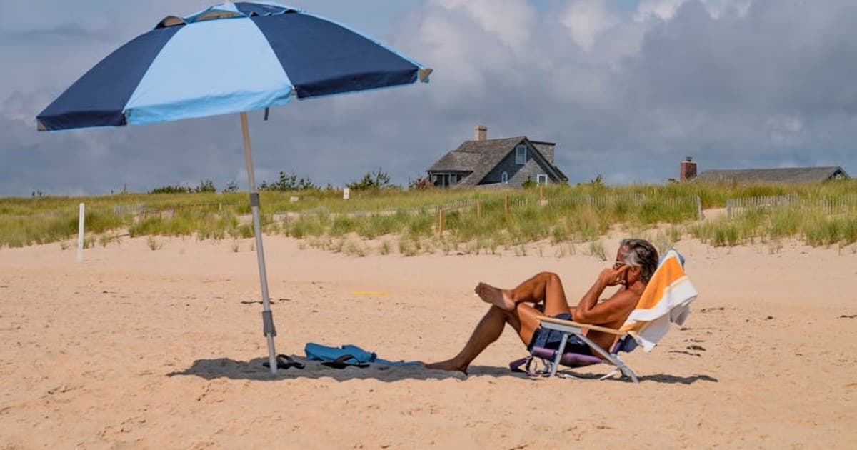 Man sunbathing under a blue umbrella on a sandy beach during summer.