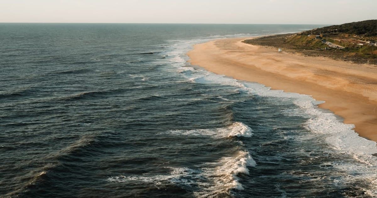 Aerial view of a tranquil beach and ocean waves on a sunny day, with golden sand and blue waters.